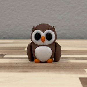 以下が含まれることがあります： 大きな黒い目をした小さな茶色のフクロウの置物。白いお腹をしています。フクロウは木製の表面に立っています。