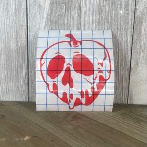 May include: A red apple sticker with a skull design. The sticker is on a white background with a blue grid pattern. The sticker is placed on a wooden surface.