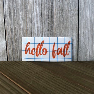 May include: A white rectangular sticker with the words "hello fall" in orange script. The sticker has a blue grid pattern and is set against a wooden background.