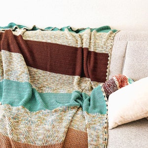 May include: A handmade crochet blanket draped over a gray sofa. The blanket features horizontal stripes in shades of brown, teal, and cream. The edges are finished with a decorative border. A pillow is visible on the sofa.