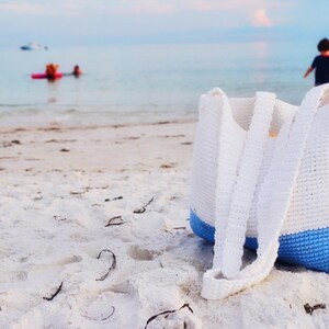 Puede incluir: Una bolsa de mano de crochet blanca con una franja azul en la parte inferior. La bolsa está sentada en una playa de arena blanca.