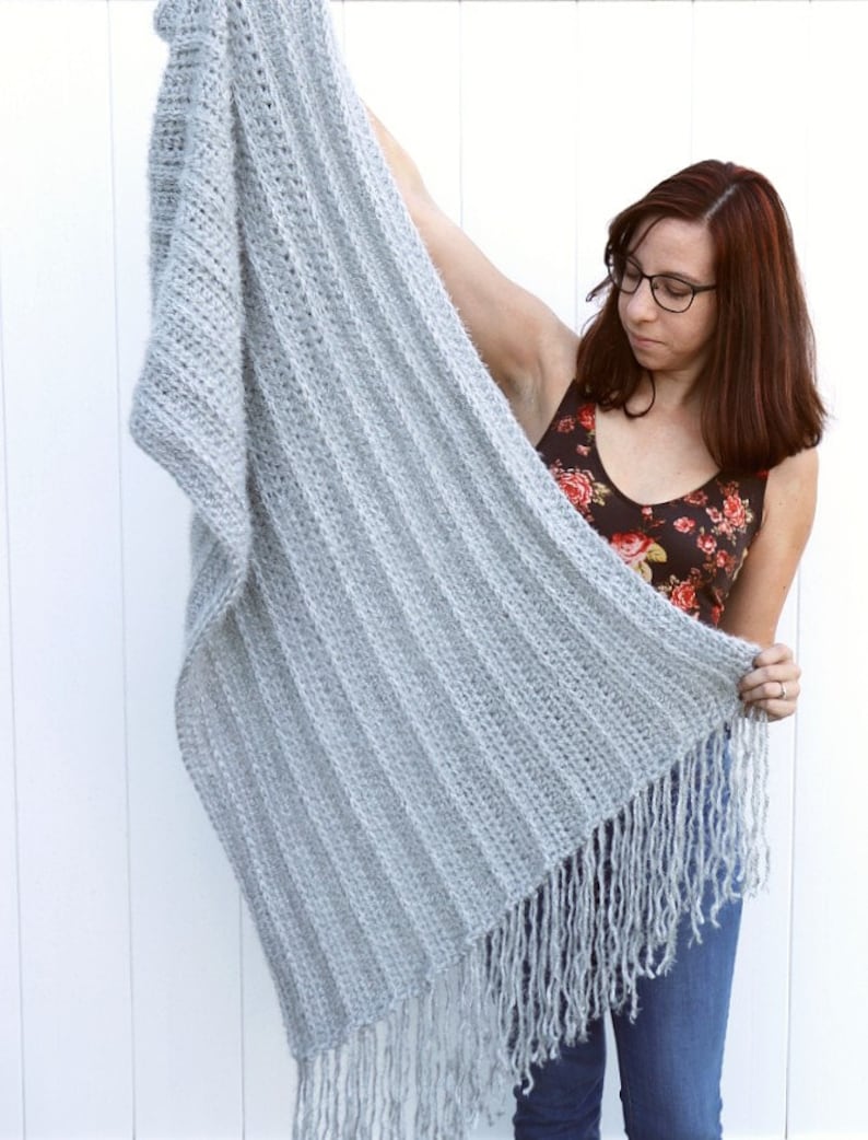 May include: A light gray crocheted throw blanket with fringe. The blanket is being held up by a person.