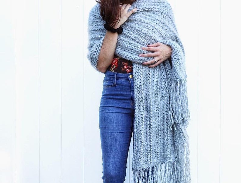 May include: A light blue crocheted shawl with fringe. The shawl is draped over a person's shoulders and is worn with blue jeans.