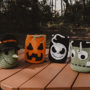 May include: Four crocheted Halloween trick-or-treat bags. The bags are shaped like a witch, a pumpkin, a skeleton, and a monster. The bags are sitting on a wooden picnic table.