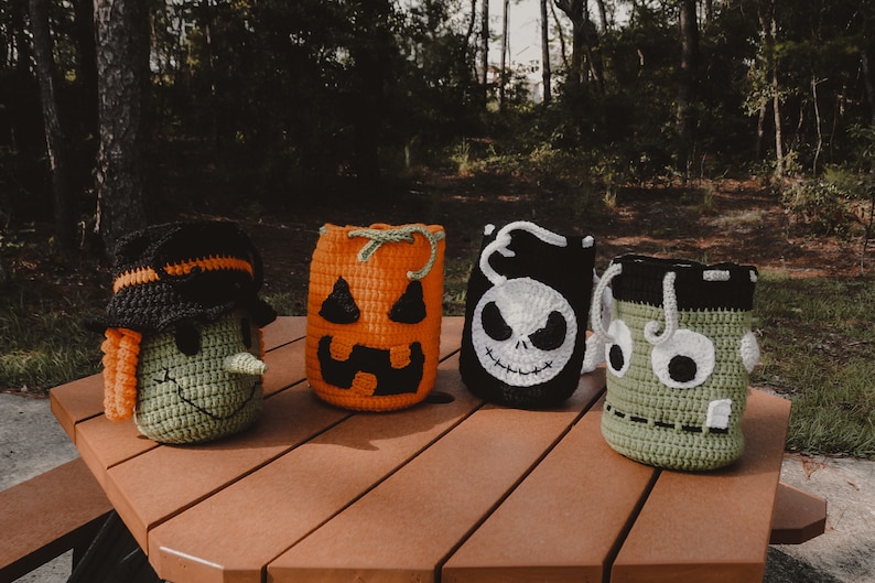 May include: Four crocheted Halloween trick-or-treat bags. The bags are shaped like a witch, a pumpkin, a skeleton, and a monster. The bags are sitting on a wooden picnic table.