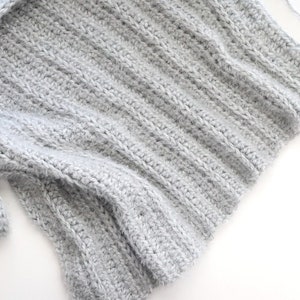 May include: A close-up of a gray crocheted fabric with a pink crochet hook lying on a white surface.