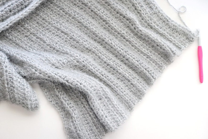 May include: A close-up of a gray crocheted fabric with a pink crochet hook lying on a white surface.