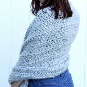 May include: A light gray crochet shawl with a fuzzy texture. The shawl is draped over a person's shoulders and is worn with blue jeans.