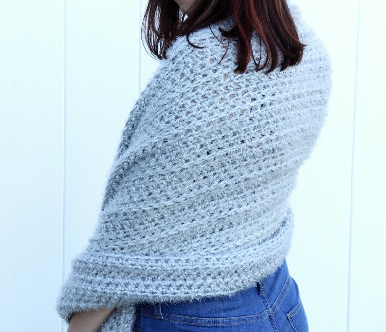 May include: A light gray crochet shawl with a fuzzy texture. The shawl is draped over a person's shoulders and is worn with blue jeans.
