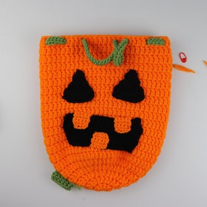 May include: A crocheted pumpkin-shaped bag with a black jack-o'-lantern face. The bag has green drawstrings and is made with orange and green yarn.