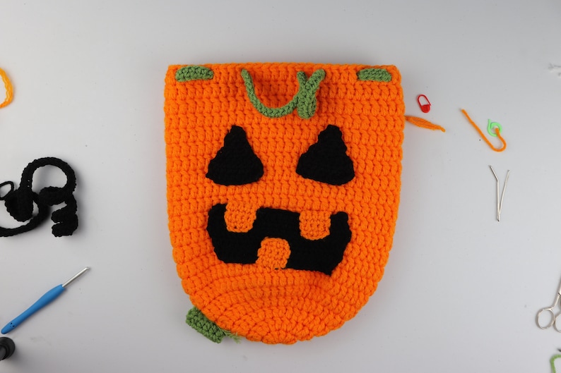 May include: A crocheted pumpkin-shaped bag with a black jack-o'-lantern face. The bag has green drawstrings and is made with orange and green yarn.