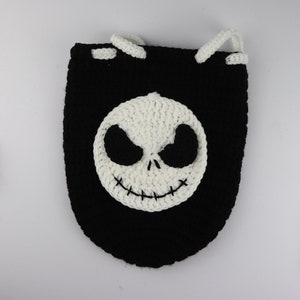 May include: A black crocheted drawstring bag with a white crocheted Jack Skellington face on the front. The bag is on a white surface with crochet hooks, yarn, and scissors.
