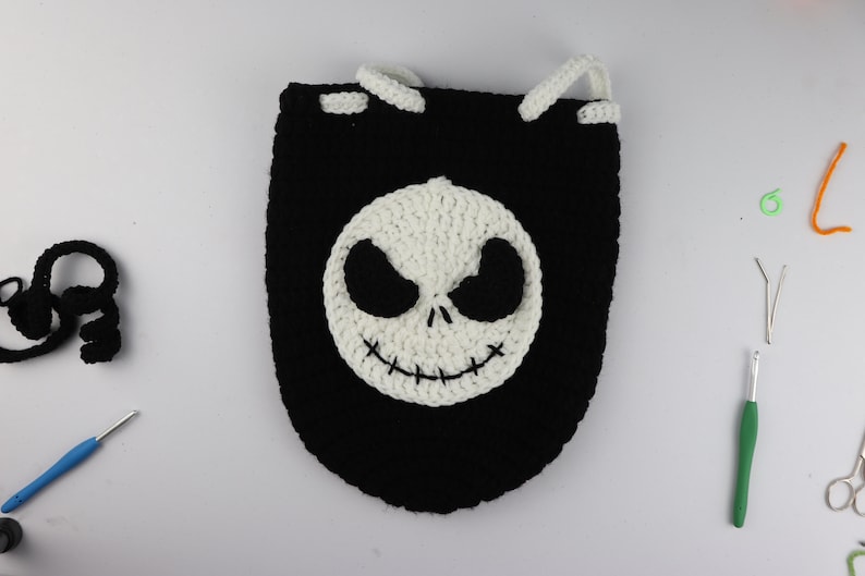 May include: A black crocheted drawstring bag with a white crocheted Jack Skellington face on the front. The bag is on a white surface with crochet hooks, yarn, and scissors.