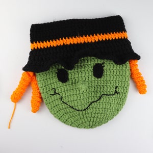 May include: Green crocheted witch face with black hat and orange yarn hair. The witch has a smiling face with black eyes and a black nose. The witch is surrounded by crafting supplies, including yarn, crochet hooks, and scissors.