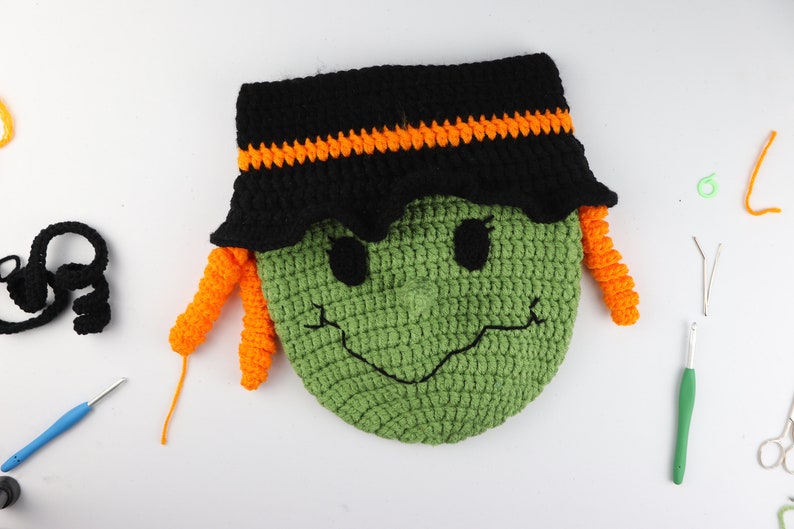 May include: Green crocheted witch face with black hat and orange yarn hair. The witch has a smiling face with black eyes and a black nose. The witch is surrounded by crafting supplies, including yarn, crochet hooks, and scissors.