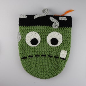 May include: Green crocheted monster bag with black eyes, white eye whites, and a stitched mouth. The bag has a black and gray crocheted top and a gray crocheted scar on the side.