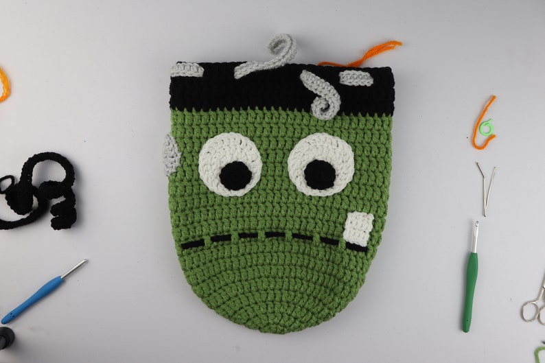 May include: Green crocheted monster bag with black eyes, white eye whites, and a stitched mouth. The bag has a black and gray crocheted top and a gray crocheted scar on the side.