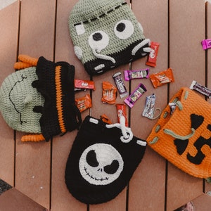 May include: Four crocheted Halloween trick-or-treat bags with different character faces. The bags are green, orange, black, and green. The bags are filled with candy bars and are sitting on a wooden table.