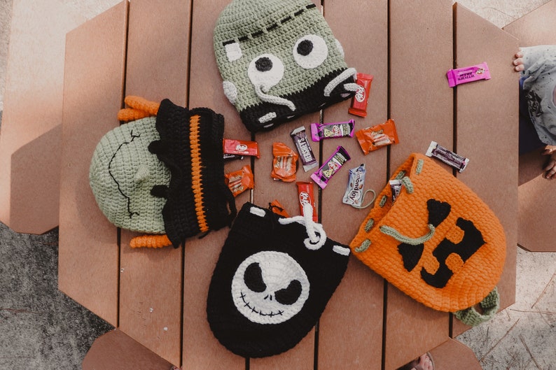 May include: Four crocheted Halloween trick-or-treat bags with different character faces. The bags are green, orange, black, and green. The bags are filled with candy bars and are sitting on a wooden table.