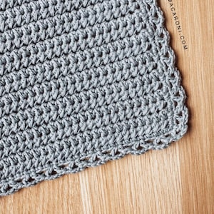 May include: Close-up of a crocheted item in a light gray color. The crochet work has a textured appearance with visible stitches. The item is placed on a wooden surface. The website address sigonimacaroni.com is visible on the right side.