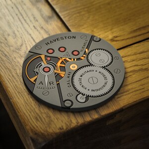 Puede incluir: Primer plano de un movimiento de reloj gris con detalles dorados. El movimiento est&aacute; etiquetado como "HAVESTON" y "CAL.H45". El texto "VINTAGE MILITARY DESIGN INDUSTRIAL DESIGN" tambi&eacute;n es visible en el movimiento.