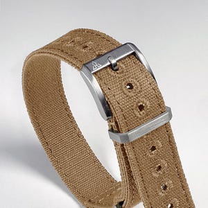 May include: Close-up of a tan canvas watch strap with a brushed silver buckle. The strap has evenly spaced holes for adjustment and a stitched edge. The buckle features a small, stylized logo.
