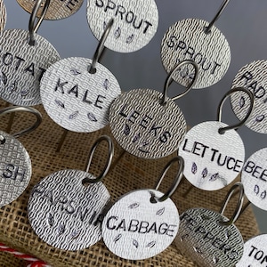 May include: A set of 14 silver metal plant markers with different vegetable names engraved on them. The markers are attached to small metal loops and have a textured surface. The names include:  'CAS', 'PROUT', 'POTAT', 'KALE', 'LEEKS', 'LETTUCE BEETROOT', 'RADISH', 'CU', 'SQUASH', 'PARSNIP', 'CABBAGE', 'TOMA', 'PEPPERS'.
