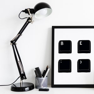 Puede incluir: Una lámpara de escritorio negra con brazo ajustable se encuentra junto a una impresión enmarcada. La impresión presenta cuatro teclas de teclado negras con las letras "B" y "E" y signos de puntuación sobre fondo blanco.