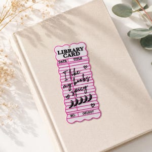Op de afbeelding: Een rechthoekige boekenlegger met geschulpte randen, een witte achtergrond en een roze rand. Het imiteert een vintage bibliotheekkaart met de tekst "LIBRARY CARD", "DATE", "TITLE", "I like my books spicy" en "NO. 061507" met chilipeper-iconen.