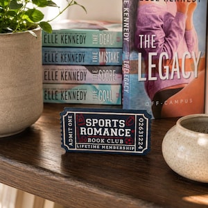 Op de afbeelding: Een donkerblauw rechthoekig bord in de vorm van een toegangsbewijs met witte tekst die Sports Romance Book Club Lifetime Membership leest, voorzien van kleine rode en witte sportpictogrammen en de woorden Admit One aan de linkerkant.