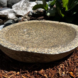 May include: A shallow, circular stone bowl featuring a speckled texture with a mix of tan, gold, and dark gray tones. The bowl has a smooth, concave interior surface and a thick, rounded rim, showcasing a natural, polished stone appearance.