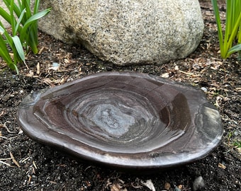 May include: A dark brown, oval-shaped stone bird bath with a smooth, polished surface. The bird bath has a shallow basin and a natural, earthy appearance. The stone has a swirling pattern of light and dark brown tones.