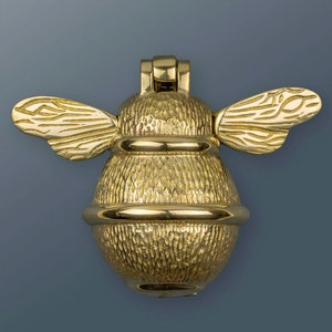 May include: A brass bee-shaped door knocker. The knocker features textured detailing on the body and wings, with a ring around the midsection. The wings are outstretched, and the knocker is mounted on a hinge.