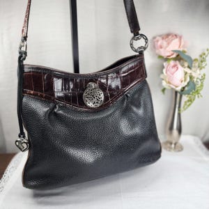 May include: Black leather shoulder bag with a brown crocodile-embossed leather trim. The bag features a silver-tone decorative medallion and a heart-shaped charm on the strap. The bag has a textured appearance and a long strap.