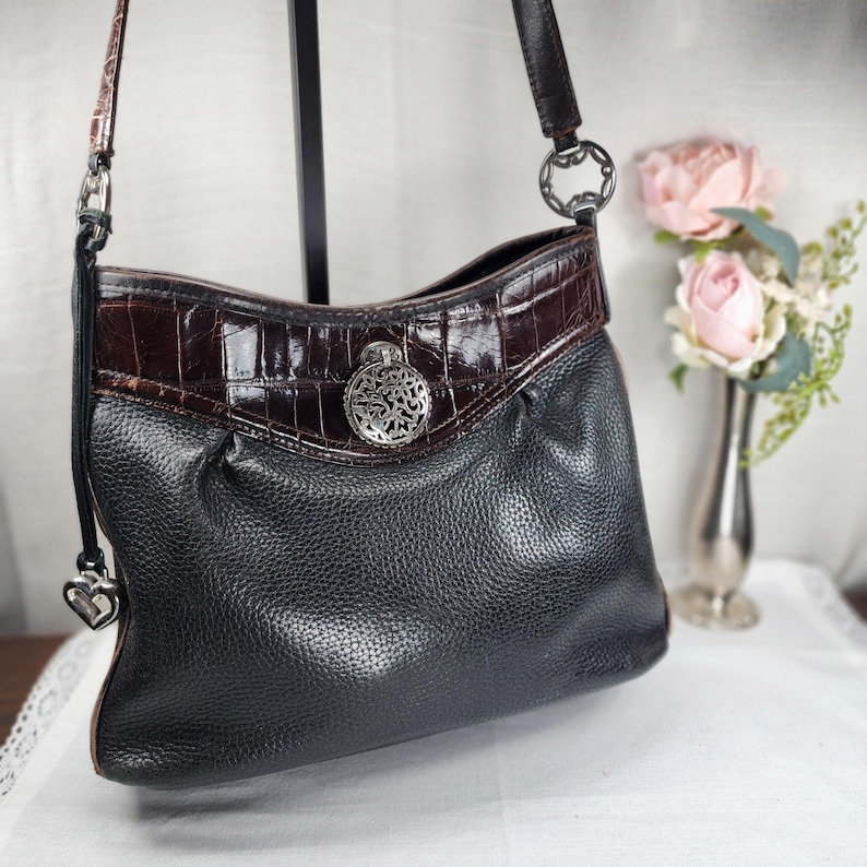 May include: Black leather shoulder bag with a brown crocodile-embossed leather trim. The bag features a silver-tone decorative medallion and a heart-shaped charm on the strap. The bag has a textured appearance and a long strap.