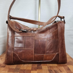 May include: Brown leather shoulder bag with a patchwork design. The bag features a long adjustable strap, a front pocket, and a metal clasp closure. The bag is made of genuine leather and has a vintage look.