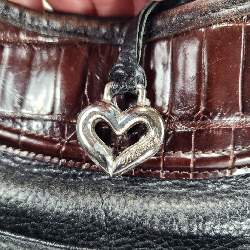 May include: Close-up of a black and brown leather handbag with a silver heart-shaped charm. The charm is engraved with the brand name "Brighton". The bag has a textured, crocodile-like pattern.