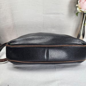 May include: Black leather shoulder bag with brown trim. The bag has a textured surface and a rounded shape. The strap is visible on the left side of the image. The bag is sitting on a white surface.