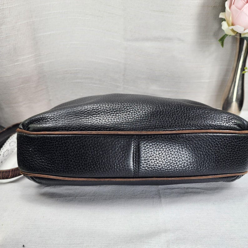May include: Black leather shoulder bag with brown trim. The bag has a textured surface and a rounded shape. The strap is visible on the left side of the image. The bag is sitting on a white surface.