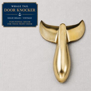 May include: A solid brass whale tail door knocker with a vintage design. The door knocker is gold and has a polished finish. The text on the sign reads "WHALE TAIL DOOR KNOCKER" and "FUNCTIONAL DECOR FOR YOUR FRONT DOOR."