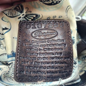 May include: Close-up of a brown leather Brighton handbag tag. The tag has embossed text that reads "D187289" and "Brighton". The tag also includes text about the handbags being crafted in China from quality leathers.