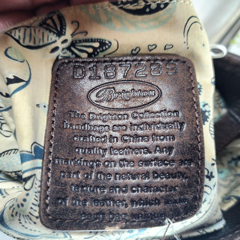 May include: Close-up of a brown leather Brighton handbag tag. The tag has embossed text that reads "D187289" and "Brighton". The tag also includes text about the handbags being crafted in China from quality leathers.