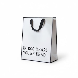 May include: White gift bag with black trim and handles. The text "IN DOG YEARS YOU'RE DEAD" is printed in black on the front of the bag. The bag is rectangular and stands upright.