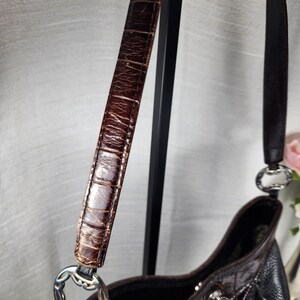 May include: A brown leather handbag with a textured, crocodile-like pattern on the strap. The bag features a silver-toned metal ring connecting the strap to the body. The bag's body is a mix of black and brown leather.