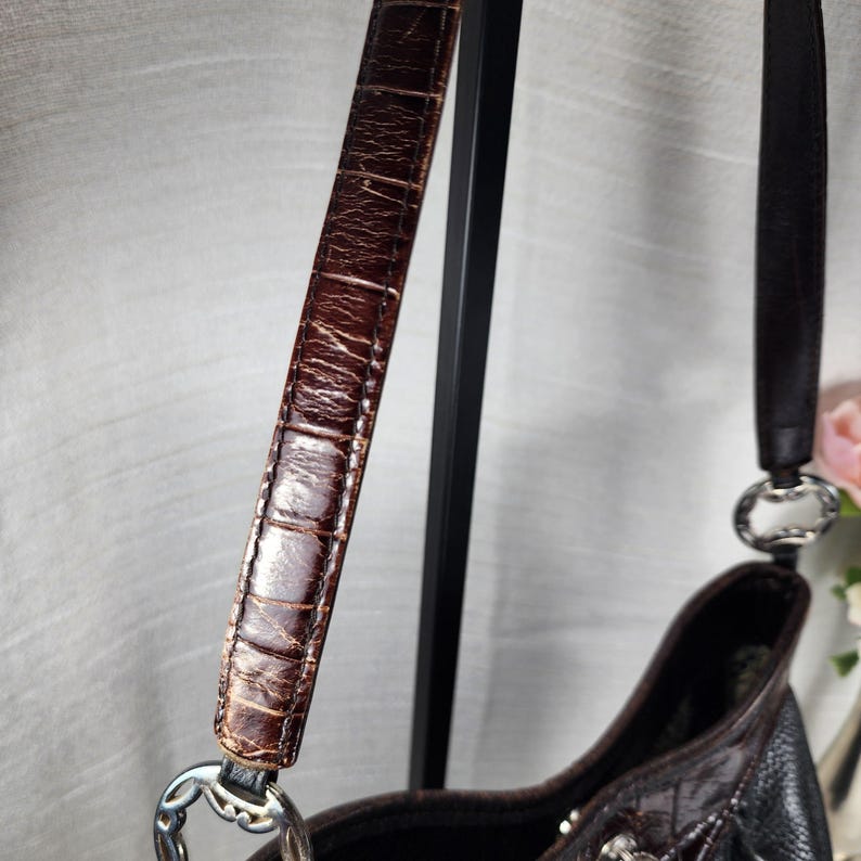 May include: A brown leather handbag with a textured, crocodile-like pattern on the strap. The bag features a silver-toned metal ring connecting the strap to the body. The bag's body is a mix of black and brown leather.