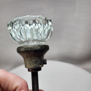 May include: Clear glass door knob with a faceted design. The knob is mounted on a tarnished metal base and rod. A small screw is visible on the side of the base. The knob is held by a hand.