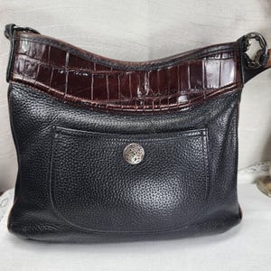 May include: Black leather shoulder bag with a brown crocodile-textured top section and a front pocket. The bag features a silver-toned decorative button on the pocket and a matching silver chain strap attachment.