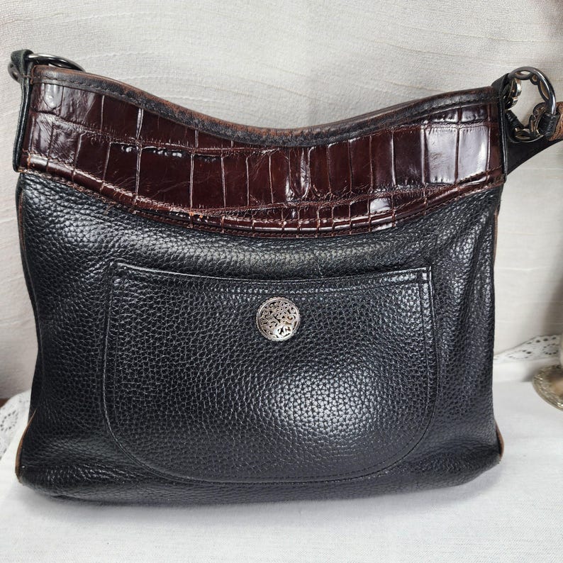 May include: Black leather shoulder bag with a brown crocodile-textured top section and a front pocket. The bag features a silver-toned decorative button on the pocket and a matching silver chain strap attachment.