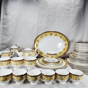 Könnte beinhalten: Ein Porzellangeschirr-Set mit weißem Grund, gelbem Blumenmuster am Rand und dunkelbrauner Umrandung. Das Set umfasst eine Teekanne, eine Zuckerdose mit Deckel, ein Milchkännchen, mehrere Teetassen, Untertassen, Speiseteller und gestapelte Teller.