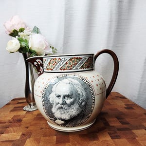 May include: Cream-colored ceramic pitcher with a portrait of a man with a long white beard. The pitcher has a brown handle and a decorative floral band with the words "Hiawatha" and "Evangeline".